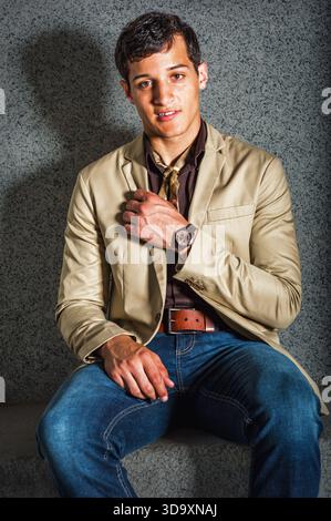 Stylish Young Man in Casual Attire. A young man sits confidently on a concrete bench. He wears a beige blazer, brown shirt, and blue jeans, accessoriz Stock Photo