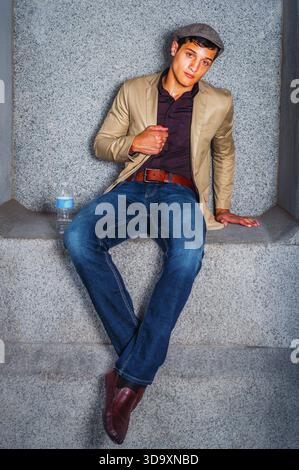 Man Sitting in Casual Attire. A young man sits casually against a stone backdrop, wearing a beige jacket and blue jeans. A bottle of water sits nearby Stock Photo