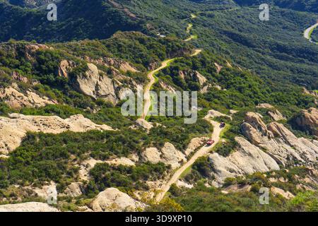 Two red off-road firefighting vehicles navigate a winding dirt road snaking through sun-drenched, rugged terrain and vibrant green chaparral in the mo Stock Photo