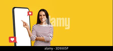 Smiling Woman Pointing at Smartphone With a Bright Yellow Background and Reaction Icons Stock Photo