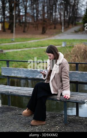 Enjoying a peaceful moment by the lakeside, a young woman in cozy winter attire engages with her mobile device while seated on a rustic bench Stock Photo