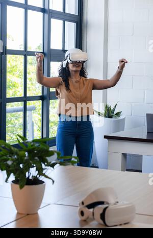 woman wearing VR headset, gesturing arms above desk with dual VR headsets and plants in office Stock Photo
