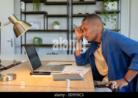 African American man resting hand on forehead while analyzing charts on laptop at desk, copy space Stock Photo
