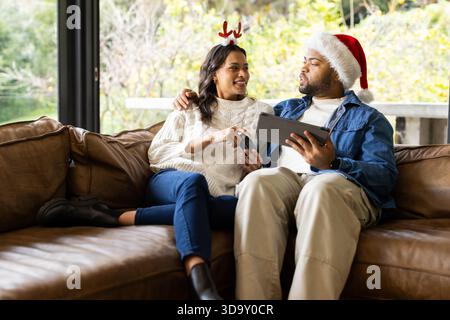 Diverse couple sitting on sofa in living room, wearing festive headbands, holding tablet and remote Stock Photo