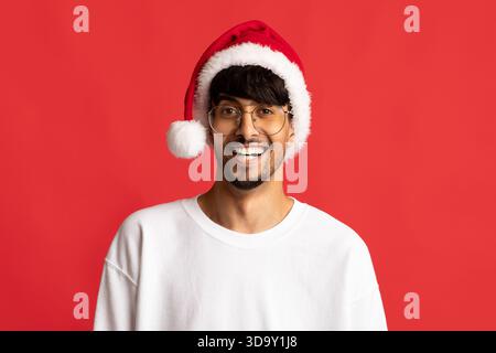 Man Smiling Cheerfully While Wearing a Christmas Hat Against a Bright Red Background During the Holiday Season Stock Photo