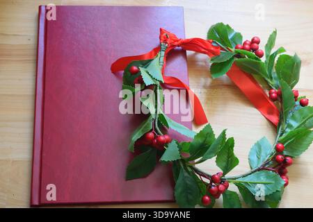 Red graduation thesis book lying on a wooden table next to a laurel wreath with green leaves, red berries, and a red ribbon, symbolizing an academic d Stock Photo