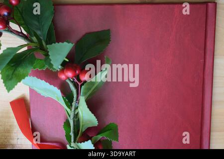 Red graduation thesis book lying on a wooden table next to a laurel wreath with green leaves, red berries, and a red ribbon, symbolizing an academic d Stock Photo