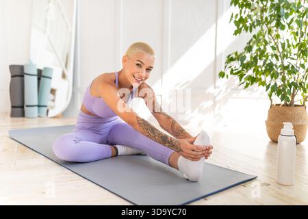 Domestic sports concept. Positive young woman in sportswear doing leg stretch exercises, sitting on mat on floor Stock Photo