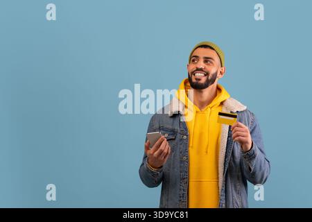 Man Wearing Yellow Hoodie and Denim Jacket Smiles While Holding Credit Card and Smartphone Against Blue Background Stock Photo