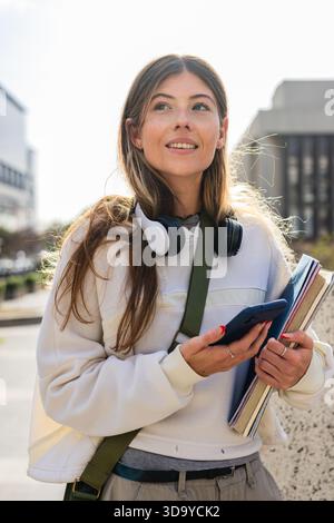 Female student standing on city walkway carrying notebooks and smartphone with headphones on neck Stock Photo