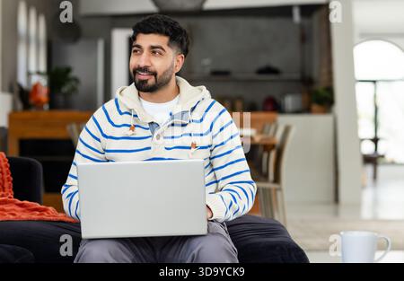 Indian man sitting on couch draped with orange throw in living area holding laptop and smiling Stock Photo