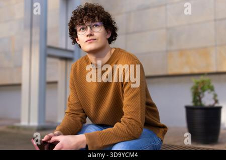 Male wearing brown sweater and glasses sitting on bench in courtyard by planter holding smartphone Stock Photo