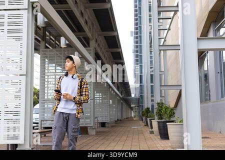 Asian man standing on urban walkway holding smartphone with backpack and headphones, copy space Stock Photo