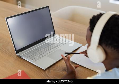 Student is writing handwritten notes with pencil in notebook at wooden table beside silver laptop Stock Photo