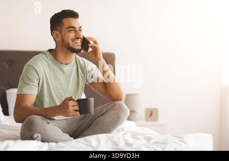 Young Man Enjoying a Phone Call While Sipping Coffee in a Cozy Bedroom Stock Photo