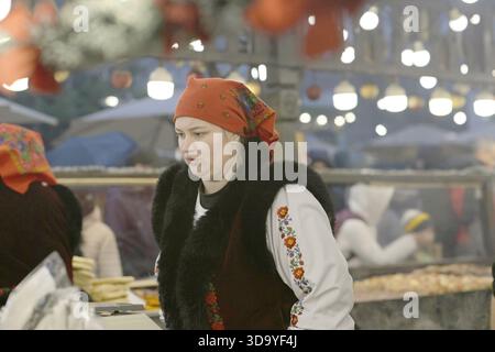 December 6, 2025 – Craiova, Romania: A food vendor wearing traditional embroidered clothing works at a steaming stall in the Christmas market as visit Stock Photo