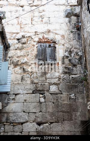 A rugged stone exterior with a worn wooden shutter over a small window. Weathered textures, moss, and exposed wires create an archaic, quiet alleyway Stock Photo