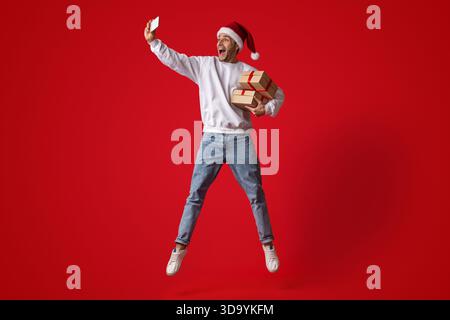 Man Joyfully Jumping With Christmas Gifts While Taking a Selfie Against a Bright Red Background Stock Photo