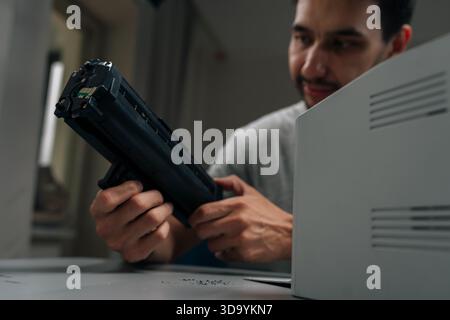 Close-up portrait of bearded repairman smiling performing printer maintenance, replacing empty toner cartridge with new one on desk, ensuring smooth Stock Photo
