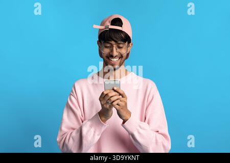 Young Man in Pink Sweater and Cap Smiles While Using Smartphone Against a Bright Blue Background Stock Photo