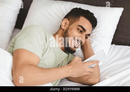 Young Man Relaxing in Bed Using Smartphone and Smiling During Morning Time Stock Photo