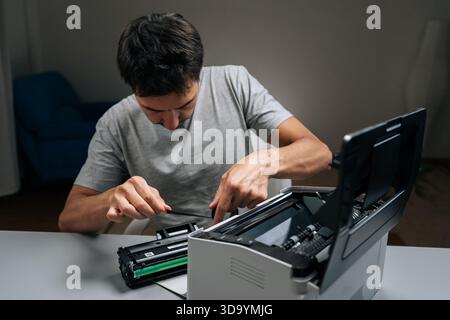 Man replacing toner cartridge and troubleshooting laser printer sitting at office desk, demonstrating maintenance and support to restore printing Stock Photo