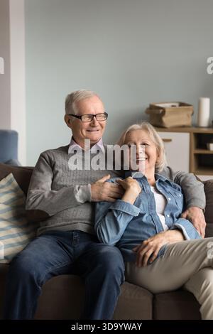 Affectionate senior husband relax on sofa embracing smiling aged wife Stock Photo