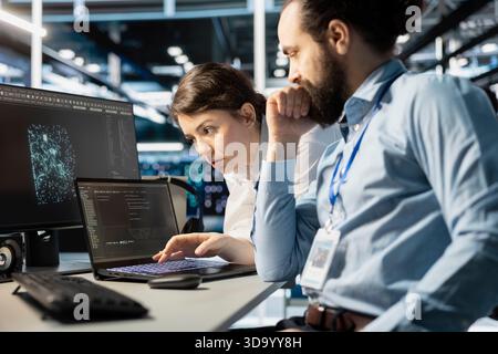 Team of programmers using notebook in server room, analyzing data. Data center office IT experts doing brainstorming, examining code on laptop, ensuring system integrity and security Stock Photo