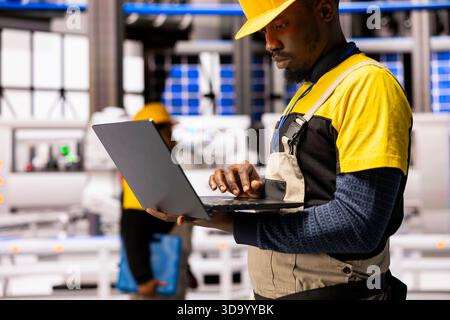 Black male engineer enhances assembly line efficiency by managing automation software on laptop. Industrial technician assesses robot arm manufacture and industry 4.0 systems in automated factory. Stock Photo