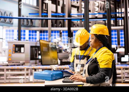 Industry 4.0 experts operating digital control panel, optimizing automation line and ensuring robotic arm accuracy. African american technicians upgrading production flow in smart manufacturing plant. Stock Photo