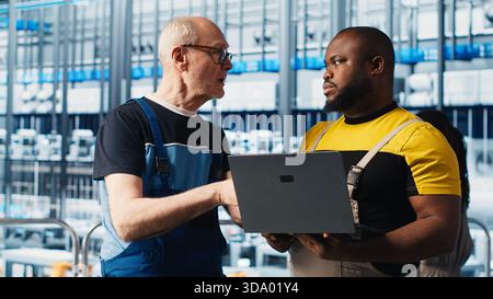 Smart factory senior engineer teaching new trainee to manage robotic arms. Industry 4.0 industrial plant team leader and intern controlling automated machinery, ensuring workflow continuity, camera A Stock Photo