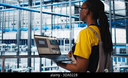 Employee using laptop in smart factory, collecting and analyzing data. African american woman using tracking software in industry 4.0 industrial plant equipped with advanced sensors, camera A Stock Photo