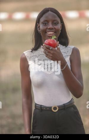 Happy, healthy young woman smiles brightly while holding fresh red apple outdoors. She is wearing a sleeveless white lace-trim top and black trousers Stock Photo