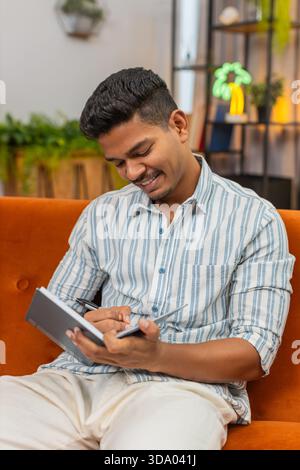 Indian man at home writes plans in notebook sets schedule and goals uses pen and keeps focus. Arabian guy on sofa checks list ticks boxes adds time notes and reviews tasks for work and study and calm. Stock Photo