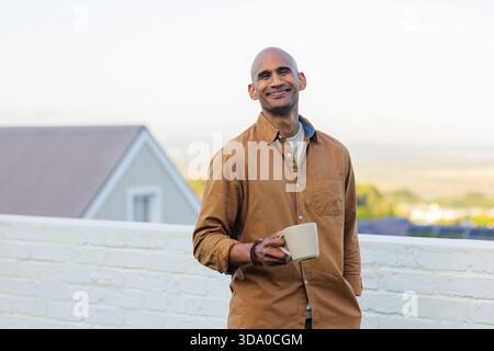 Man standing on white brick rooftop terrace railing holding ceramic mug overlooking green fields Stock Photo