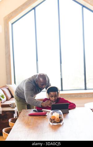 African American father and son leaning over table in living room, using tablet with headphones Stock Photo