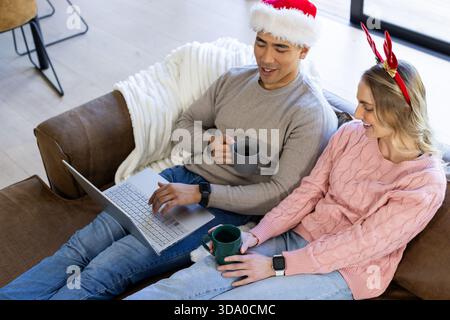 Diverse couple wearing Santa hat and antlers, sitting on couch at home holding mugs, using laptop Stock Photo