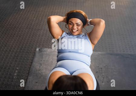 Mid-adult woman doing sit-ups on textured mat at gym wearing blue workout set, smartwatch, hoops Stock Photo
