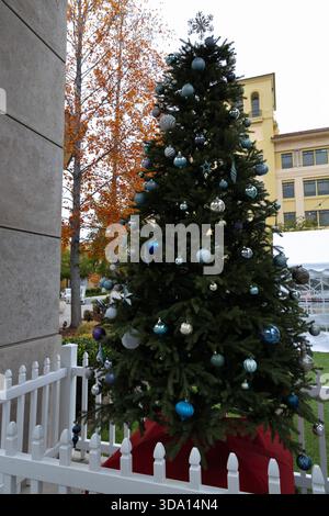 A Christmas tree is set up in a park area with various ornaments. Nearby, autumn leaves can be seen on trees. Stock Photo