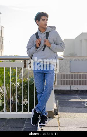 African American man leaning against metal railing on sunlit rooftop terrace with backpack straps Stock Photo