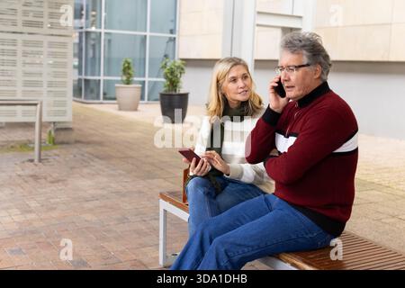 Senior couple sitting on bench in courtyard, woman holding smartphone and man talking on phone Stock Photo