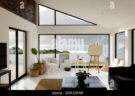 Blank canvas standing on wooden easel in coastal art studio overlooking ocean through glass doors Stock Photo