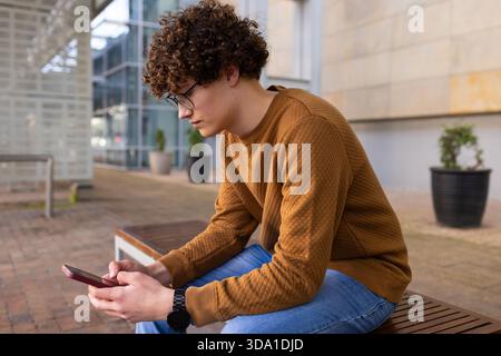 Man in his twenties sitting on wooden bench near planters in plaza using smartphone, copy space Stock Photo
