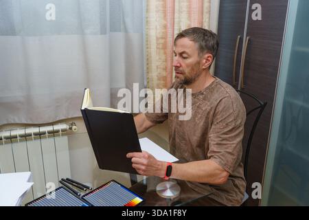 A focused middle-aged man sits at a glass desk, examining an open black sketchbook. His workspace includes colored pencils and blank paper for drawing Stock Photo