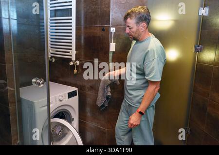 A middle-aged man in a light green sweatsuit holds a towel, preparing to do laundry in a modern bathroom with a front-loading washing machine. Stock Photo
