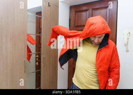 A bearded man puts on a bright orange hooded jacket in a home hallway. He stands by a mirrored wardrobe, getting ready to go out. Stock Photo