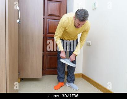 A middle-aged Caucasian man with a beard, wearing a yellow long-sleeved shirt and mismatched orange and grey socks, bends over to put on a grey slip-o Stock Photo