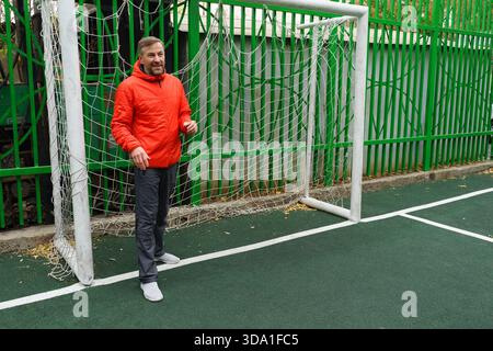 A smiling middle-aged man in a bright orange jacket stands inside a soccer goal on an outdoor sports court. The court has green artificial turf and wh Stock Photo