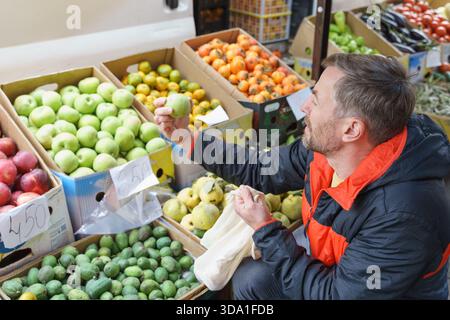 A middle-aged man with a beard shops for fresh fruit at an outdoor farmers market. He is inspecting a green apple while holding a reusable mesh bag, w Stock Photo