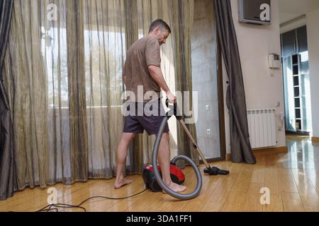 A barefoot middle-aged man in casual clothes is vacuuming the polished wooden floor of his modern apartment during the day. Stock Photo